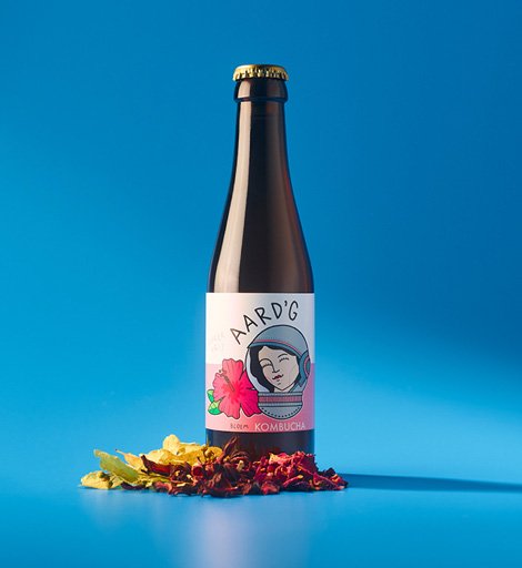 Een fles Aard'g Bloem Kombucha op een blauwe achtergrond met de ingrediënten hibiscus, linde en rozenbottel ernaast.