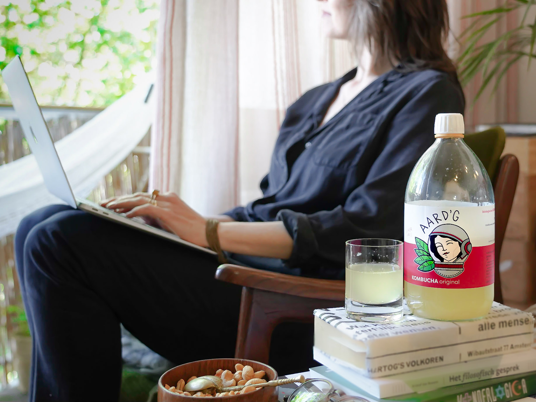 Een fles Aard'g Kombucha Original op een stapel boeken met op de achtergrond een vrouw die naar buiten kijkt met een laptop op haar schoot.