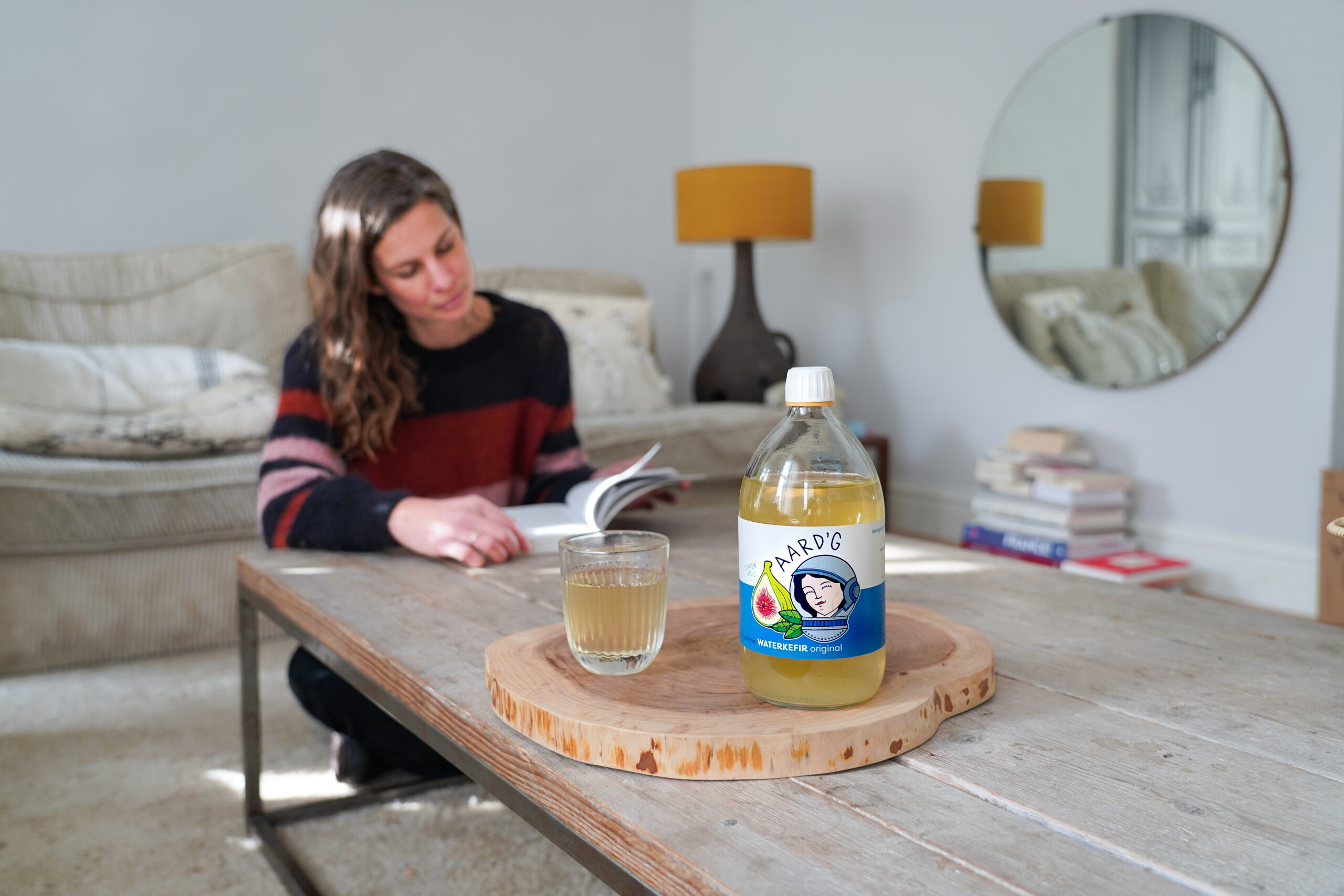 Een fles waterkefir original van Aard'g staat op een tafel op een houten plankje. Een vrouw leest een magazine aan dezelfde tafel.